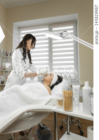 Aesthetician in white uniform applies facial care procedure to a relaxed female client in a bright, contemporary spa room. Beauty products and equipment create a clean, professional atmosphere Aesthetician in white uniform applies facial care procedure to a relaxed female client in a bright, contemporary spa room. Beauty products and equipment create a clean, professional atmosphere 143829987