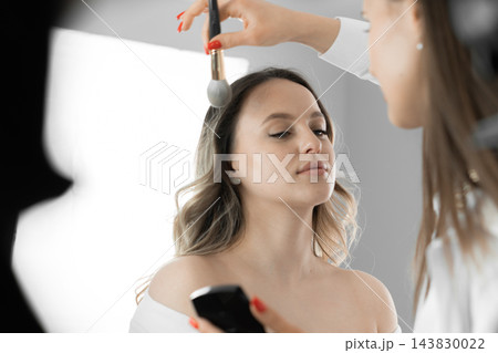 A makeup artist uses a brush to enhance a woman's complexion in a well lit studio environment. The scene captures the elegance and precision of professional beauty services in action 143830022