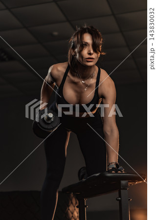 Confident woman working out with a dumbbell, demonstrating proper form and dedication in a dimly lit fitness center. Strength training and healthy lifestyle concept for motivation and empowerment Confident woman working out with a dumbbell, demonstrating proper form and dedication in a dimly lit fitness center. Strength training and healthy lifestyle concept for motivation and empowerment 143830032