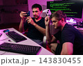 Focused two men with controllers sit at computer with programming code on screen. Dark room with purple and pink neon lights. Concept for gaming, development, esports, technology, teamwork. 143830455