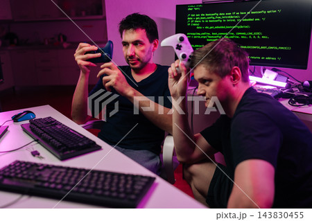Focused two men with controllers sit at computer with programming code on screen. Dark room with purple and pink neon lights. Concept for gaming, development, esports, technology, teamwork. 143830455