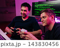 Positive two young men enjoy playing video games in dark room with neon light. One smiling holding controller as friend watches. Computer with code in background. Concept of fun, friendship, hobby. 143830456