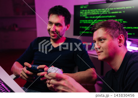 Positive two young men enjoy playing video games in dark room with neon light. One smiling holding controller as friend watches. Computer with code in background. Concept of fun, friendship, hobby. Positive two young men enjoy playing video games in dark room with neon light. One smiling holding controller as friend watches. Computer with code in background. Concept of fun, friendship, hobby. 143830456