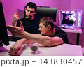 Two male colleagues working together, discussing project on screen. Intense focus and collaboration in modern office with powerful gaming computer and vibrant pink neon lighting. 143830457