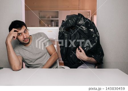 Humorous scene of puzzled young man looking at camera anonymous person sitting nearby with black trash bag over their head. Absurd concept concept of confusion, strange relationships, hiding. Humorous scene of puzzled young man looking at camera anonymous person sitting nearby with black trash bag over their head. Absurd concept concept of confusion, strange relationships, hiding. 143830459