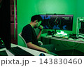 Pensive man sitting at desk in dark room, illuminated green light, looking stressed, focused on computer screen showing map and data. Concept for cybersecurity, hacking, programming, intense gaming. 143830460