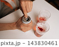 Close-up of male hand opening chilled aluminum beverage can on white table, preparing to pour drink into two glasses filled with foamy red liquid, indicating moment of relaxation. 143830461