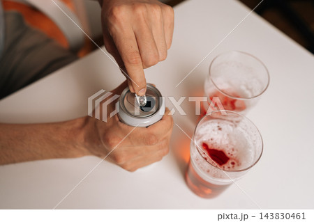 Close-up of male hand opening chilled aluminum beverage can on white table, preparing to pour drink into two glasses filled with foamy red liquid, indicating moment of relaxation. Close-up of male hand opening chilled aluminum beverage can on white table, preparing to pour drink into two glasses filled with foamy red liquid, indicating moment of relaxation. 143830461