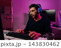 Serious focused male gamer in headphones intensely playing on pc, typing on keyboard in cyberpunk style dark room lit by neon pink lights, streaming and focused on competition. 143830462