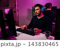 Two young men playing competitive online computer game in dark interior of room, focusing intently on their screens wearing headsets and interacting with their keyboards. 143830465