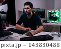 Serious young man concentrates on monitor screen, working or gaming late. High-tech setup includes powerful computer with green lights and multiple screens. Dark room with intense mood. 143830488
