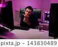 Man wearing headphones sitting at desk with multiple monitors and sends air kiss. Playful gamer or streamer interacting with audience in purple neon light. Modern tech setup for online activity. 143830489