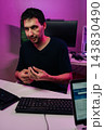 Vertical portrait of male professional working on computer setup in modern home office, handling small device, with keyboards and monitors visible under ambient neon lighting. 143830490