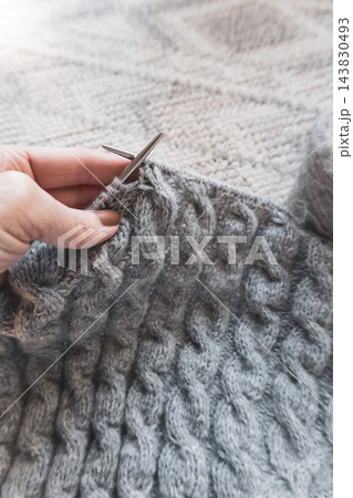 Hands knitting gray cable garment with metal needles. Close-up view of the textured stitches and knitting in progress. Cable sweater on needles. Gray wool knitted sweater. Cozy autumn hobby. 143830493