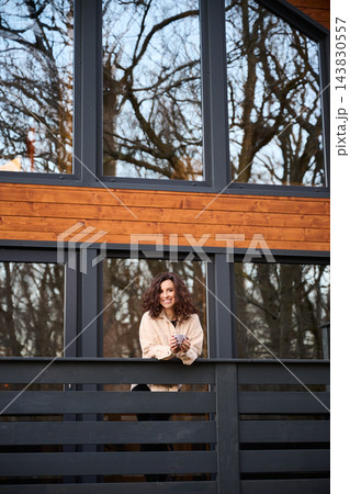 Smiling woman standing on balcony of modern wooden house, representing lifestyle, comfort, real estate and contemporary living environment Smiling woman standing on balcony of modern wooden house, representing lifestyle, comfort, real estate and contemporary living environment 143830557