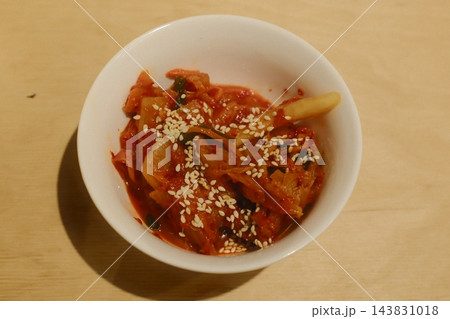 Vibrant red Korean kimchi, sesame-sprinkled, in ceramic bowl on light wood indoors Vibrant red Korean kimchi, sesame-sprinkled, in ceramic bowl on light wood indoors 143831018