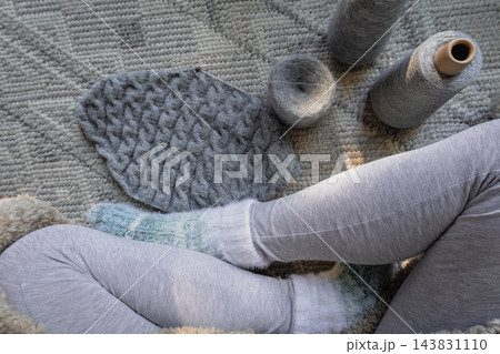 Gray cable garment with yarn balls near crossed legs in knitted socks. Cable sweater on needles with woman legs on carpet. Cozy autumn hobby. Hygge lifestyle. Comfortable domestic life. Gray cable garment with yarn balls near crossed legs in knitted socks. Cable sweater on needles with woman legs on carpet. Cozy autumn hobby. Hygge lifestyle. Comfortable domestic life. 143831110