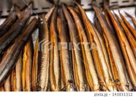 Rows of golden smoked eels hang from metal rack at traditional market, showing glossy skin and rich texture. Close-up seafood display highlights artisanal smoking, preservation and regional cuisine Rows of golden smoked eels hang from metal rack at traditional market, showing glossy skin and rich texture. Close-up seafood display highlights artisanal smoking, preservation and regional cuisine 143831312