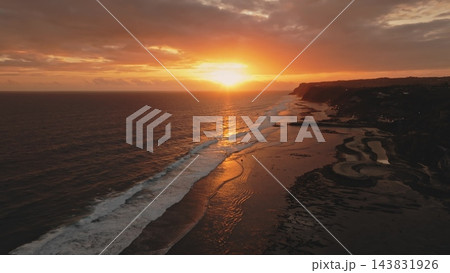 Golden sunset over the tranquil ocean at Melasti Beach, Bali, creating a vibrant orange reflection on the water and wet sand as white waves roll onto the tropical coastline. Aerial view drone flight Golden sunset over the tranquil ocean at Melasti Beach, Bali, creating a vibrant orange reflection on the water and wet sand as white waves roll onto the tropical coastline. Aerial view drone flight 143831926
