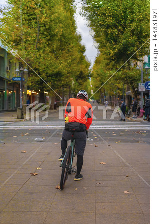 Cyclist in City Street. Cyclist in orange jacket and helmet waits with bicycle on pedestrian street lined with green trees. Cyclist faces road crossing, outdoor sporting activity in urban surroundings Cyclist in City Street. Cyclist in orange jacket and helmet waits with bicycle on pedestrian street lined with green trees. Cyclist faces road crossing, outdoor sporting activity in urban surroundings 143831971