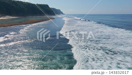 Indonesia, Bali island, Bukit. Aerial view tracking ocean waves breaking on coral reef near a tropical sand beach with green cliffs and clear blue water, creating white foam and sparkling sunlight Indonesia, Bali island, Bukit. Aerial view tracking ocean waves breaking on coral reef near a tropical sand beach with green cliffs and clear blue water, creating white foam and sparkling sunlight 143832017