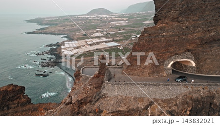 Tenerife, Mirador Punta del Fraile. Dramatic coastal road carved into cliffs with a natural stone tunnel, winding along volcanic coastline above the Atlantic Ocean. Aerial view drone flight panorama 143832021