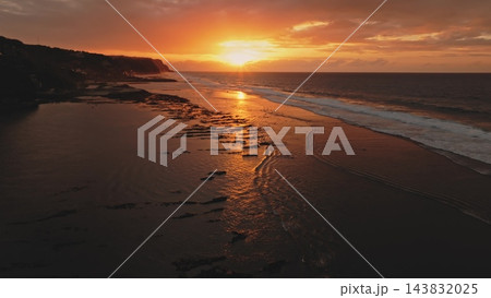 Melasti Beach in Bali, Indonesia, showcasing a warm golden hour sunset with sun reflections on the wet sand and breaking ocean waves along the picturesque coastline and dramatic cliffs 143832025