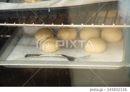 Bread dough balls rising on parchment inside oven with metal fork on tray, warm bakery scene showing proofing process, soft light, rustic home baking and preparation 143832138