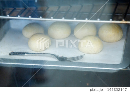 Fresh dough balls rising on parchment lined baking tray inside warm oven with metal fork in foreground, soft steam haze, home baking preparation, simple kitchen food process 143832147