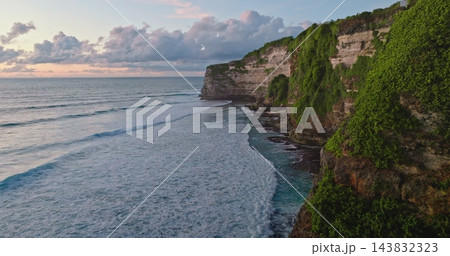 Tropical Bali coastline aerial view of dramatic limestone cliffs draped in lush green vegetation meeting turquoise Indian Ocean with gentle surf and pastel evening sky 143832323