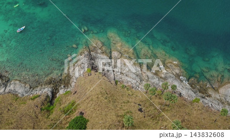 Tropical island coastline, rocky cliffs meeting the clear turquoise sea, boats anchored in the shallow waters and dry grass covering the hillside under a bright sky. Aerial view top down drone shot Tropical island coastline, rocky cliffs meeting the clear turquoise sea, boats anchored in the shallow waters and dry grass covering the hillside under a bright sky. Aerial view top down drone shot 143832608