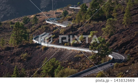 Tenerife, Canary Islands: Aerial view capturing cars riding winding road through Tenerife's volcanic terrain, with lush pine trees contrasting dark rock. Static shot Tenerife, Canary Islands: Aerial view capturing cars riding winding road through Tenerife's volcanic terrain, with lush pine trees contrasting dark rock. Static shot 143832669