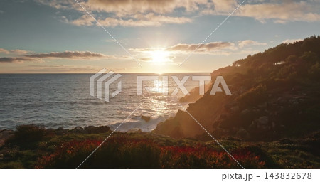 Sunset warm light over rugged coastline of Portugal, illuminating ocean waves hitting the cliffs and vibrant vegetation on rocky shore. Golden hour sun casting beautiful nature landscape. Drone flight 143832678