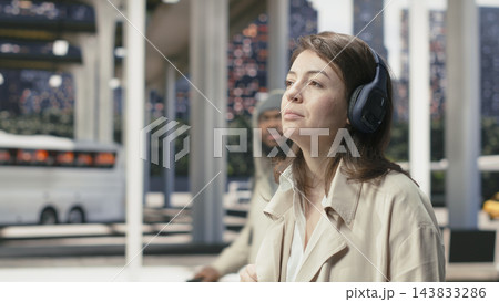 Close up of relaxed woman wearing headphones while walking to bus terminal, listening to music while waiting for departure. Traveler enjoying songs inside coach station 143833286