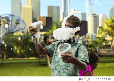 African child with virtual reality headset standing on picnic blanket and holding controllers as parents relax beside food and basket in sunny urban park. VR interactive technology. 143833356