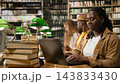 Student, laptop and keyboard lead as woman in tan jacket types at shared library desk beside stacked books. Digital learning, academic research and study skills support independent coursework. 143833430