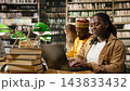 Student, laptop and books frame woman typing at wooden library table with classmates studying behind her. Coursework, digital literacy and academic preparation support learning through technology. 143833432