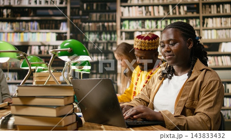 Student, laptop and books frame woman typing at wooden library table with classmates studying behind her. Coursework, digital literacy and academic preparation support learning through technology. Student, laptop and books frame woman typing at wooden library table with classmates studying behind her. Coursework, digital literacy and academic preparation support learning through technology. 143833432