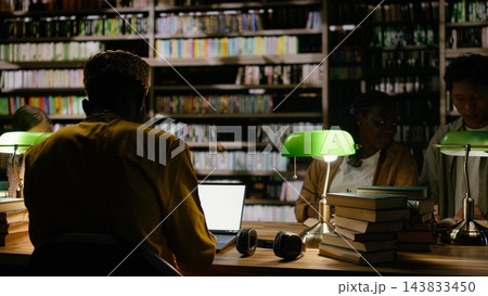 Adult learner, laptop and library frame rear view of man in yellow clothing working at computer near open books. Continuing education, research and digital skills shape study at communal table. 143833450