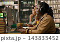 Student, computer and headphones as smiling woman types at desktop workstation beside other learners. Digital learning, concentration and productivity support research at library desks. 143833452