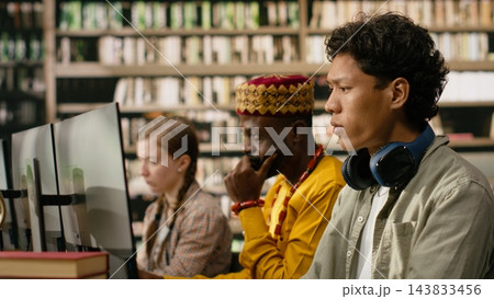 Student, headphones and computer as young man focuses on desktop monitor while classmates work nearby in library. Digital literacy, independent study and academic focus support computer learning. 143833456