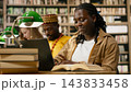 Student, laptop and book as smiling woman works on keyboard beside open reference text at library table. Digital learning, productivity and research connect academic reading with computer coursework. 143833458