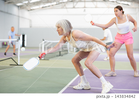 Elderly woman bouncing ball in pickleball game on court with racket 143834842