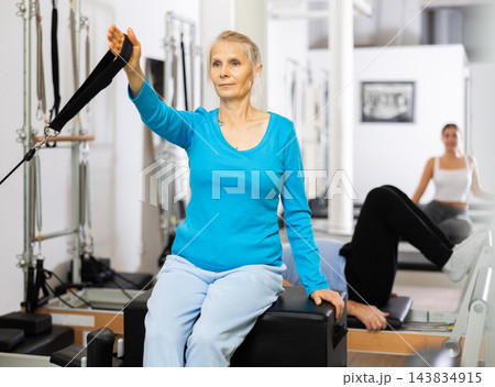 Elderly woman holding hand in adjustable sling during workout on Pilates reformer combo bed machine in gym Elderly woman holding hand in adjustable sling during workout on Pilates reformer combo bed machine in gym 143834915