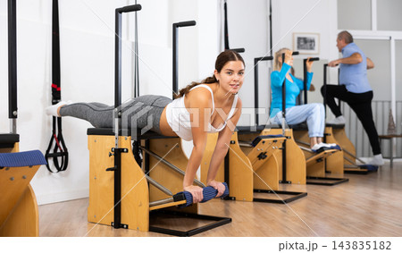 Positive young female training hands and lower back while laying on Pilates Wanda chair in fitness studio 143835182