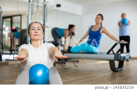 Group of young elderly people in sportswear exercising Pilates with mini balls while lying on mats in rehabilitation center Group of young elderly people in sportswear exercising Pilates with mini balls while lying on mats in rehabilitation center 143835219