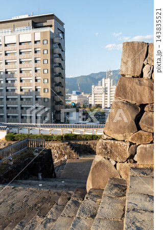 石垣と石段越しに見る、高層ホテル風の建物と市街地・山並みc 143835521