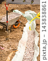 A wheelbarrow loaded with gray gravel stands next to a freshly dug trench lined with protective fabric at a busy landscaping site. 143838861