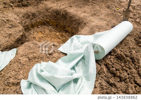 Gardening preparations are in progress with unrolled landscape fabric positioned next to a freshly dug hole, indicating a site ready for planting in sandy soil. 143838862