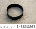 Under soft lighting, a black rubber drive belt rests flat on a gray concrete background. This mechanical component is crucial for machinery operation and shows a textured surface. 143838863